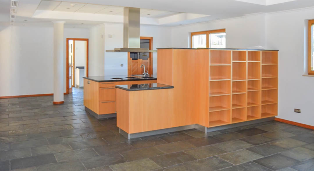 Cocina de concepto abierto con suelos de pizarra, encimeras de granito negro y mobiliario de madera clara. Incluye una isla central con campana de acero y estanterías integradas.