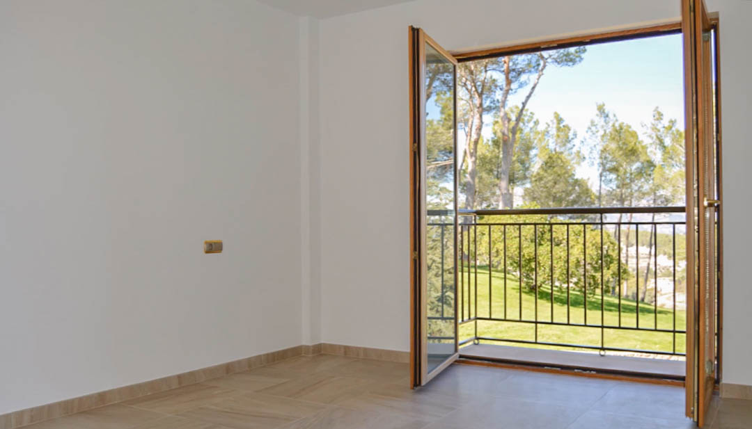 Habitación luminosa con suelos cerámicos y puertas de madera de alta calidad que dan acceso a un balcón privado con vistas al entorno natural y jardines.