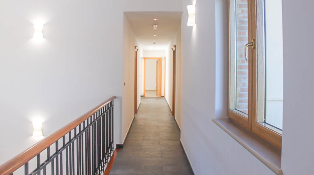 Pasillo de la planta superior con suelo de baldosas oscuras, barandilla de madera y metal, y apliques de pared modernos que iluminan el acceso a los dormitorios.