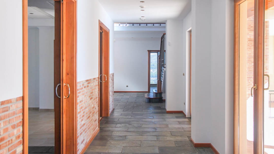 Pasillo interior con suelo de piedra natural, zócalos de ladrillo visto y carpintería de madera maciza. La estancia cuenta con iluminación empotrada y acceso a la escalera principal.
