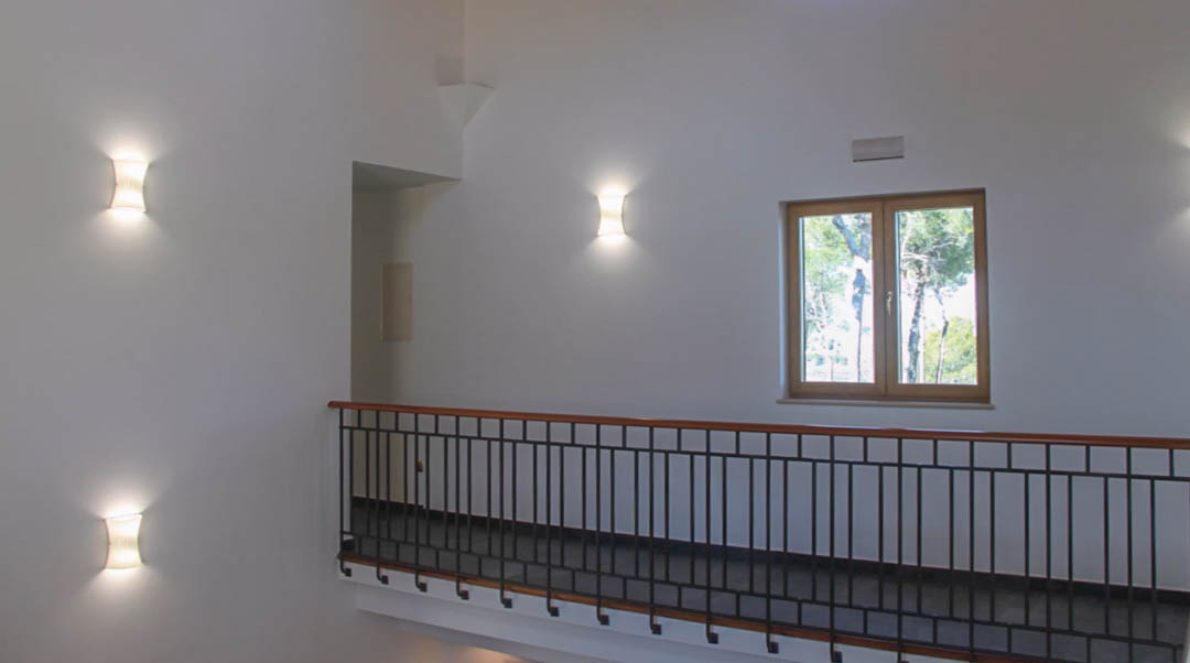 Pasillo superior con barandilla de hierro y madera, iluminación mediante apliques de pared y ventana con marco de madera que ofrece vistas al entorno natural.