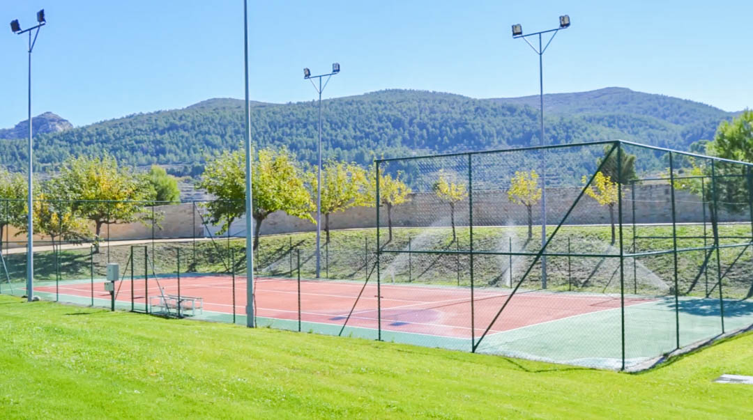 Pista de tenis vallada con superficie rojiza y sistema de iluminación profesional, rodeada de zonas verdes y vistas despejadas a las montañas.
