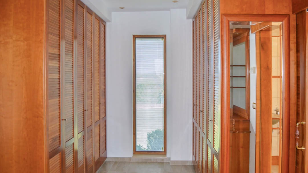 Vestidor con amplios armarios empotrados de madera de alta calidad y puertas de lamas. Una ventana vertical aporta luz natural al pasillo con suelos cerámicos y focos empotrados.