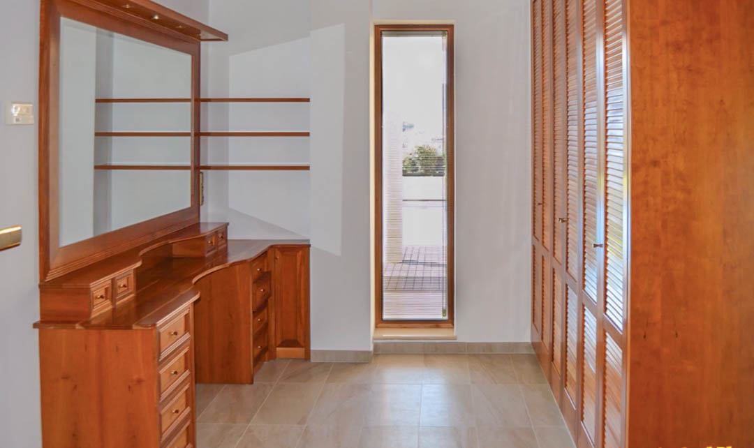 Vestidor con tocador de madera de alta calidad, armarios empotrados con puertas de persiana y ventana vertical que aporta luz natural.