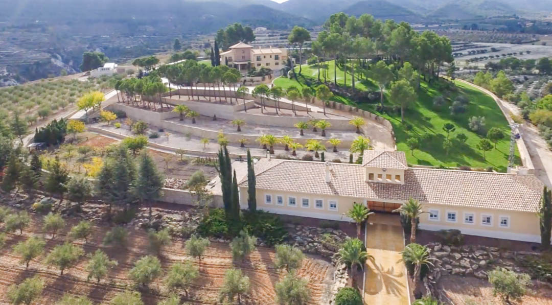 Vista aérea de la finca mostrando el edificio de almacén en primer plano, jardines aterrazados con palmeras y la villa principal rodeada de pinos y montañas.