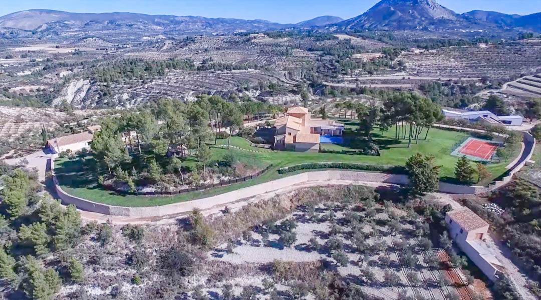 Vista aérea de la finca que muestra la villa principal, la piscina, la pista de tenis y los jardines rodeados por montañas y campos de cultivo aterrazados.