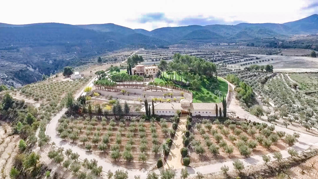 Vista aérea de la propiedad que destaca la villa principal y el almacén, rodeados de extensos campos de olivos y almendros bajo un entorno de valles y montañas.