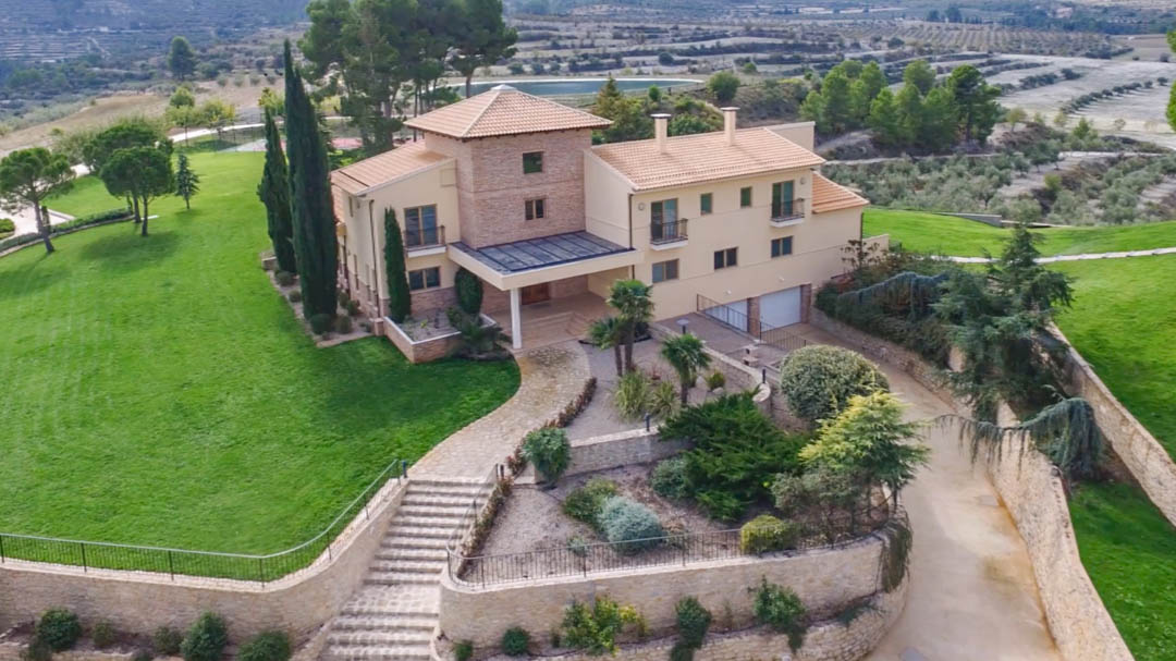 Vista aérea de la villa principal con fachada de piedra y yeso, rodeada de jardines cuidados, muros de piedra natural y vistas a los valles y campos de cultivo.