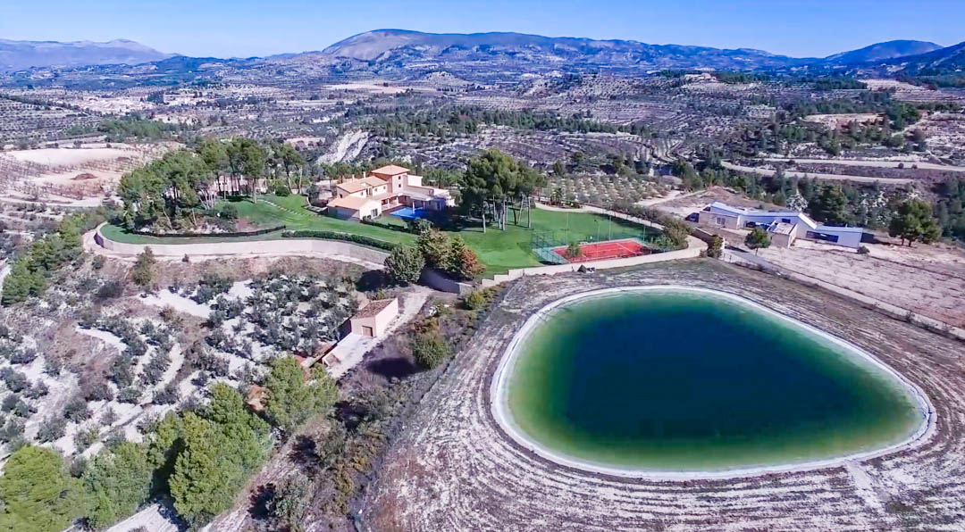 Vista aérea panorámica que integra el gran embalse de agua, la pista de tenis roja y la villa principal, rodeados de extensos campos y relieve montañoso.