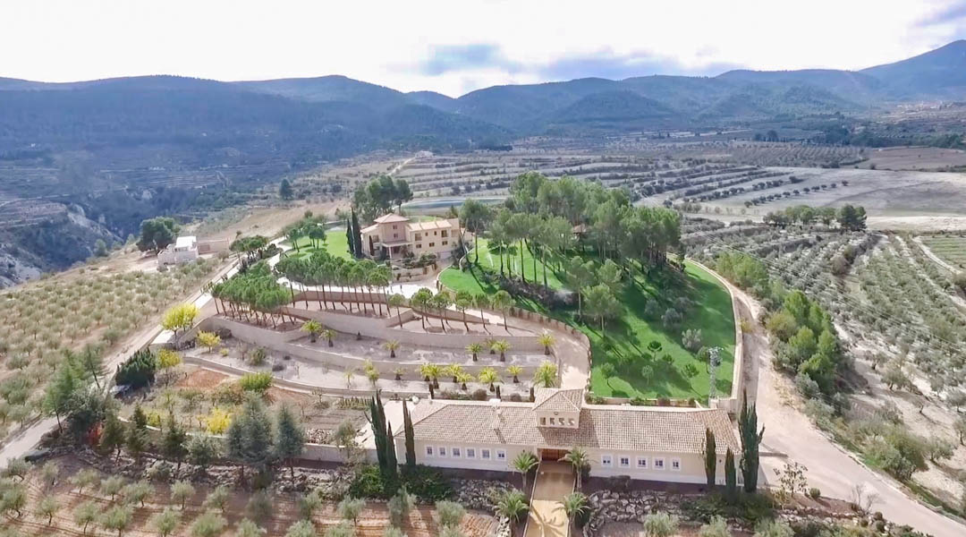 Vista aérea panorámica que muestra la villa principal y el edificio anexo, rodeados de jardines aterrazados con palmeras, pinos y extensas plantaciones agrícolas entre montañas.