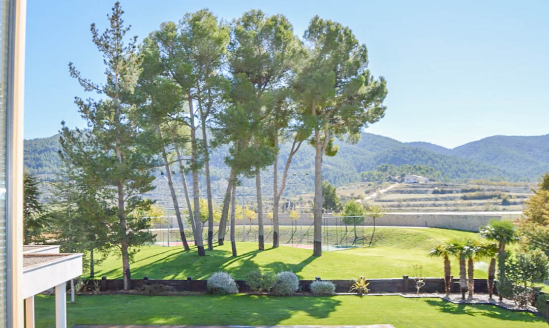 Vista de los jardines con césped cuidado, pinos maduros y una pista de tenis privada, enmarcada por montañas y campos aterrazados bajo un cielo despejado.