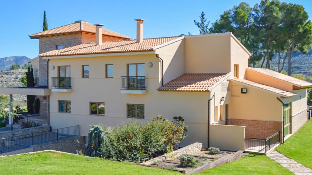 Vista exterior de la villa principal con fachada en tonos crema, tejado de teja cerámica y balcones de forja, rodeada de jardines y vistas a la montaña.