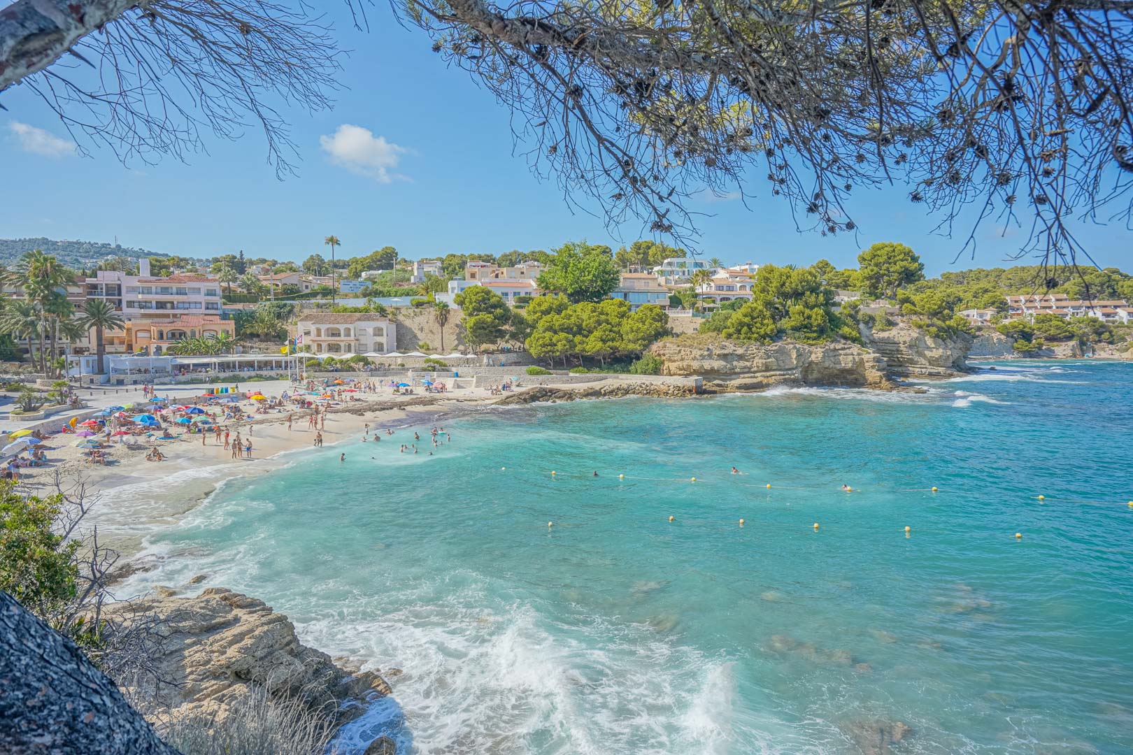 Vista panorámica de una cala de aguas turquesas en Benissa, rodeada de acantilados bajos, pinos mediterráneos y edificaciones costeras bajo un cielo despejado.