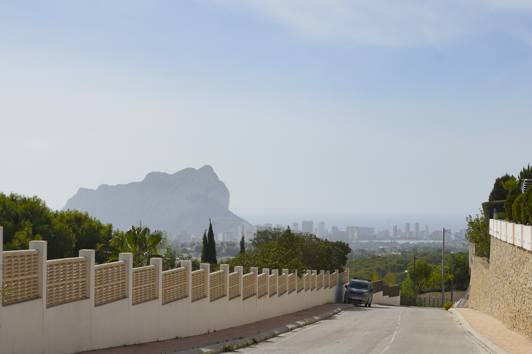 Vista panorámica desde la calle de acceso hacia el Peñón de Ifach y el skyline de Calpe, enmarcada por muros de piedra natural y celosías blancas.