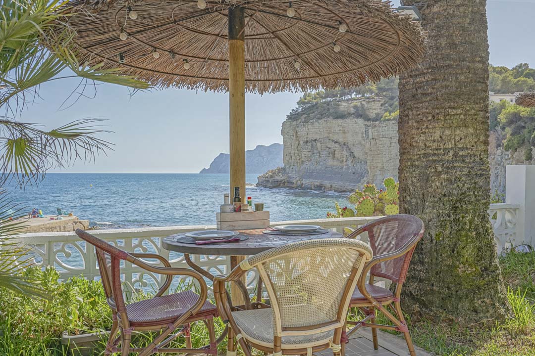 Terraza exterior con mesa de comedor bajo una sombrilla de brezo, ofreciendo vistas frontales al mar Mediterráneo y a los acantilados de la costa de Benissa.