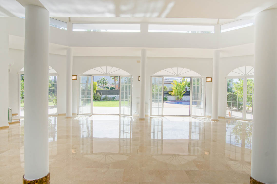 Amplio salón de estilo colonial con columnas circulares, suelos de mármol pulido y múltiples puertas francesas arqueadas que conectan con el jardín y la piscina.