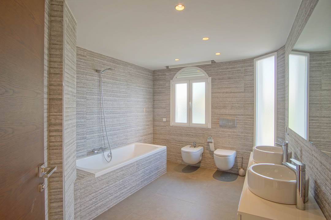 Baño moderno con doble lavabo de diseño, bañera integrada y revestimiento cerámico texturizado. Incluye sanitarios suspendidos y luz natural a través de una ventana de arco.