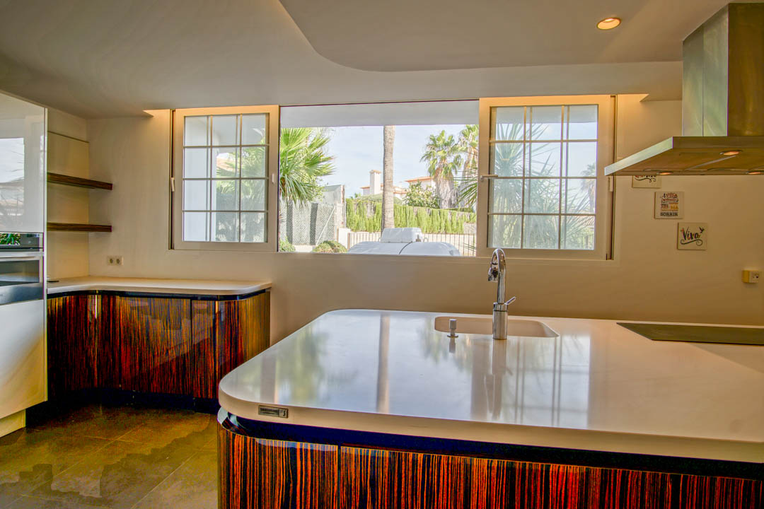 Cocina moderna con isla blanca, armarios de diseño curvo en madera oscura y amplios ventanales que ofrecen luz natural y vistas al jardín.