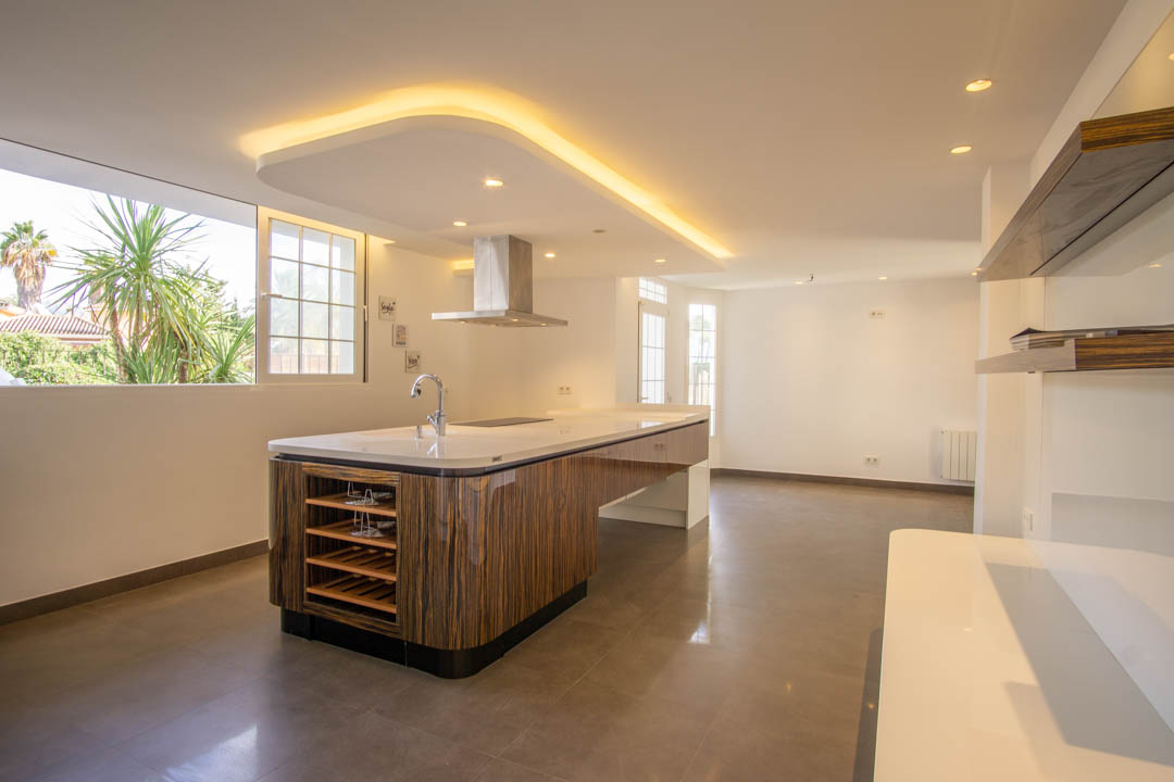 Cocina moderna con isla central de madera veteada, encimera blanca y campana de acero. Destaca el techo con iluminación LED indirecta y las vistas al jardín mediterráneo.