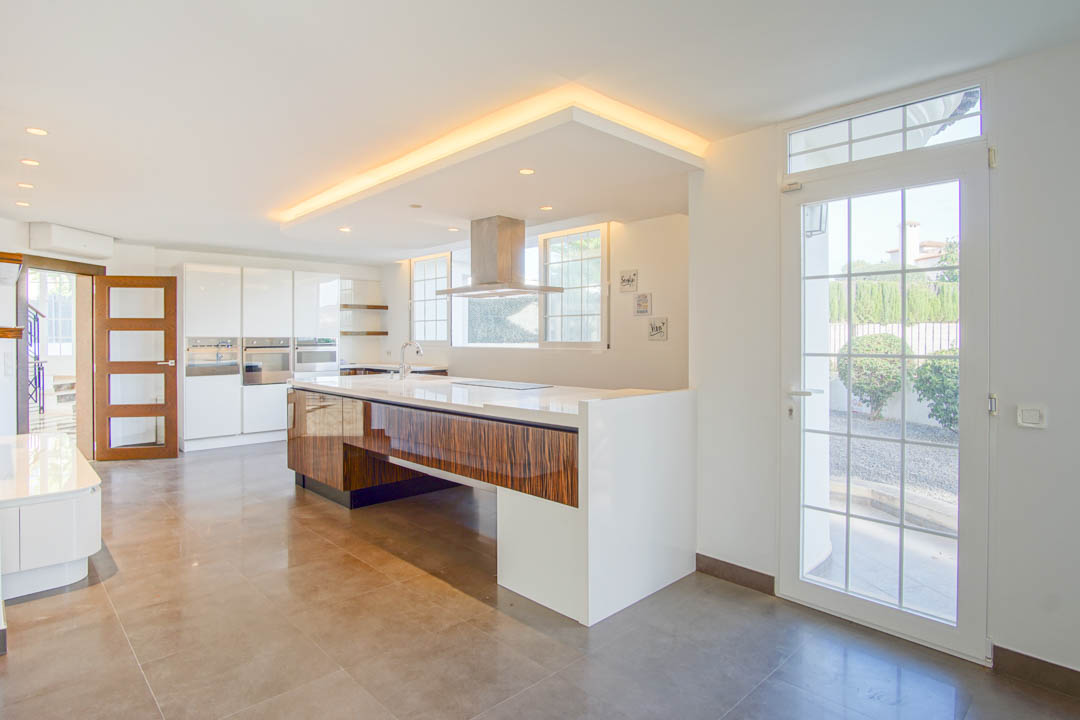 Cocina moderna con isla central de madera veteada, encimera blanca y campana de acero. Incluye iluminación perimetral en el techo y acceso directo al exterior.
