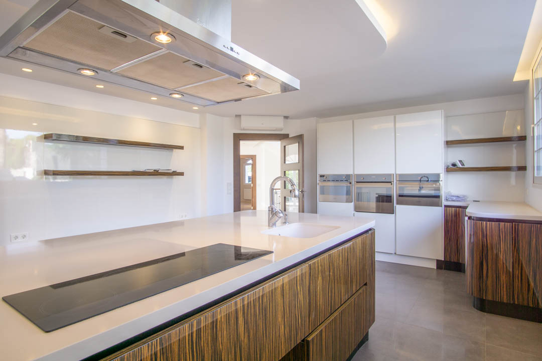 Cocina moderna con isla central, encimeras blancas y armarios de madera veteada. Incluye electrodomésticos integrados, campana de acero y estantes flotantes minimalistas.
