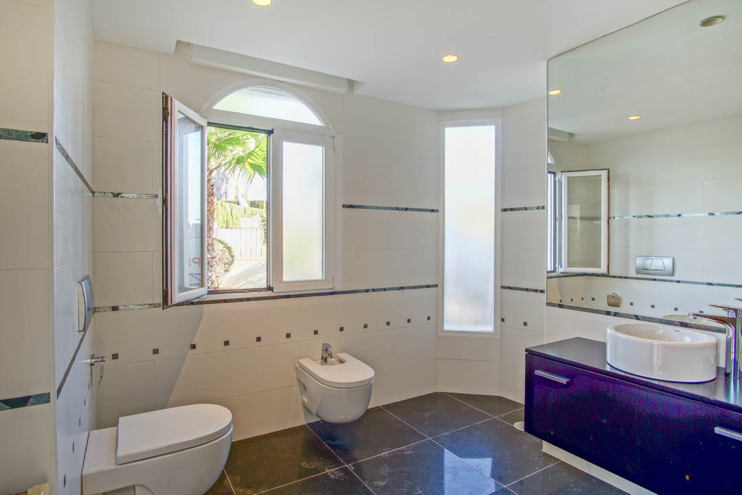 Cuarto de baño con sanitarios suspendidos, lavabo sobre encimera oscura y azulejos blancos con detalles decorativos. Ventana de arco con vistas a las palmeras exteriores.