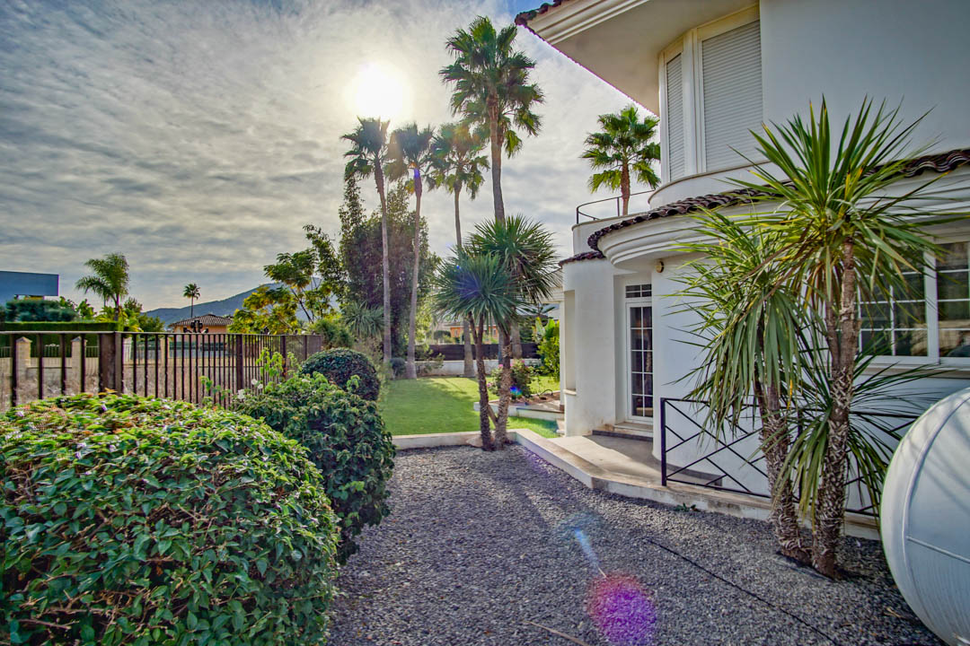Fachada blanca de estilo colonial con líneas curvas, rodeada de un jardín con palmeras, arbustos cuidados y senderos de grava.