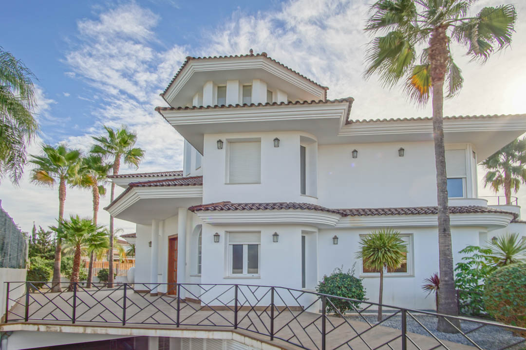 Fachada de estilo colonial con paredes blancas, tejados de teja cerámica y barandillas de hierro forjado negro, rodeada de palmeras maduras.