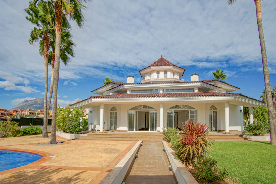 Fachada exterior de estilo colonial con porche de columnas blancas, torre central octogonal y jardín con palmeras y piscina bajo un cielo despejado.