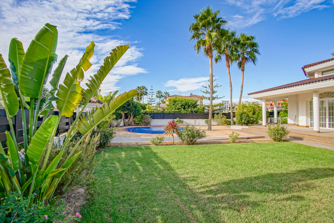 Jardín maduro con césped impecable, palmeras altas y piscina privada circular, junto a un porche cubierto con columnas de estilo colonial.