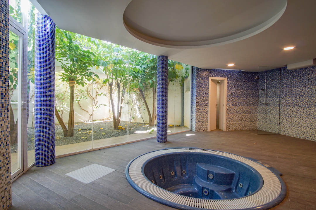 Sala de spa con jacuzzi circular encastrado, columnas de mosaico azul y amplios ventanales con vistas directas a un patio inglés privado con vegetación.