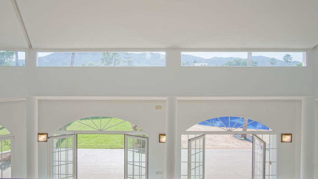 Salón de doble altura con ventanales de arco hacia el jardín y la piscina, destacando las ventanas superiores con vistas despejadas a las montañas.
