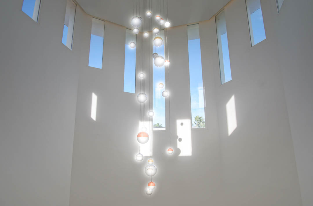 Techo de doble altura con una moderna lámpara de esferas de cristal suspendidas y múltiples ventanas verticales que aportan abundante luz natural al recibidor.
