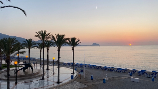Vista del amanecer desde el paseo marítimo cercano, con palmeras, una escultura de ancla y la playa equipada con sombrillas frente al mar Mediterráneo.