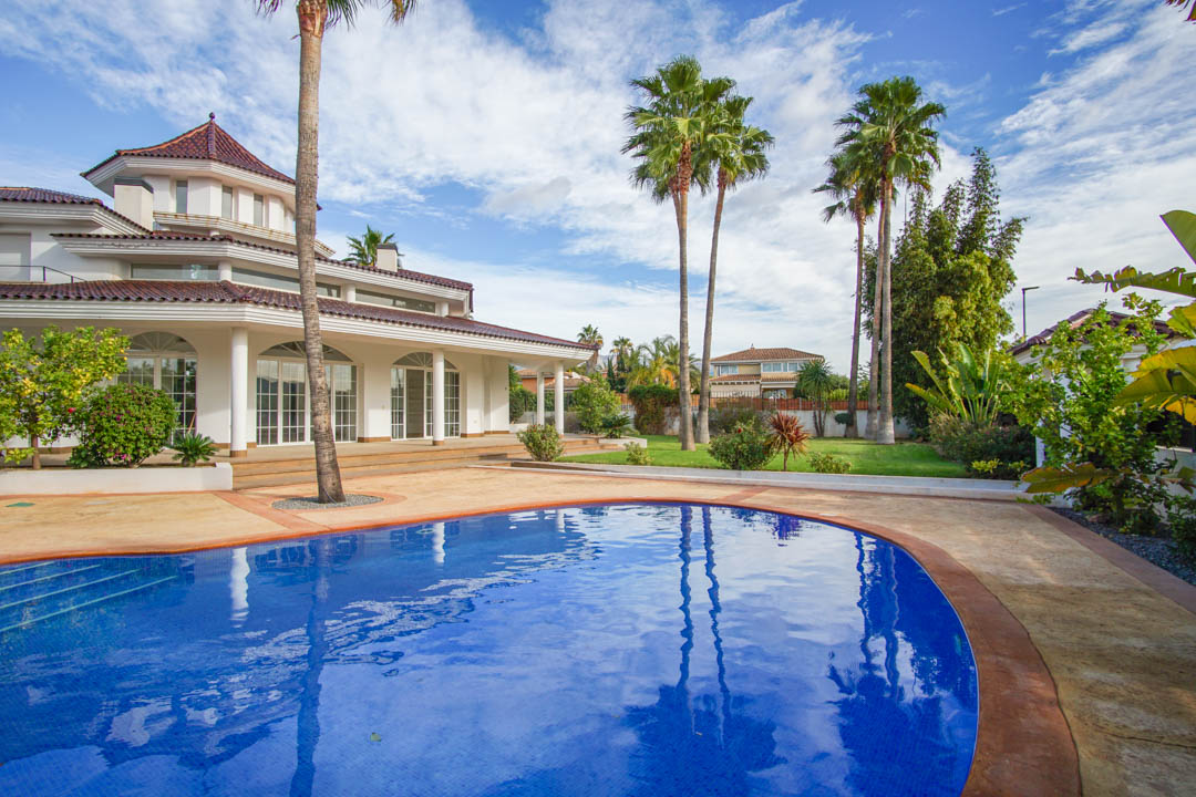 Vista exterior del chalet colonial con piscina curva, jardín de palmeras maduras y fachada blanca con amplios ventanales y columnas.