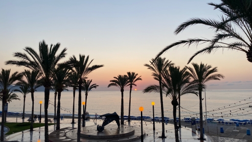 Vistas panorámicas del amanecer sobre el paseo marítimo, con palmeras maduras, iluminación ambiental y una escultura central frente al mar Mediterráneo.