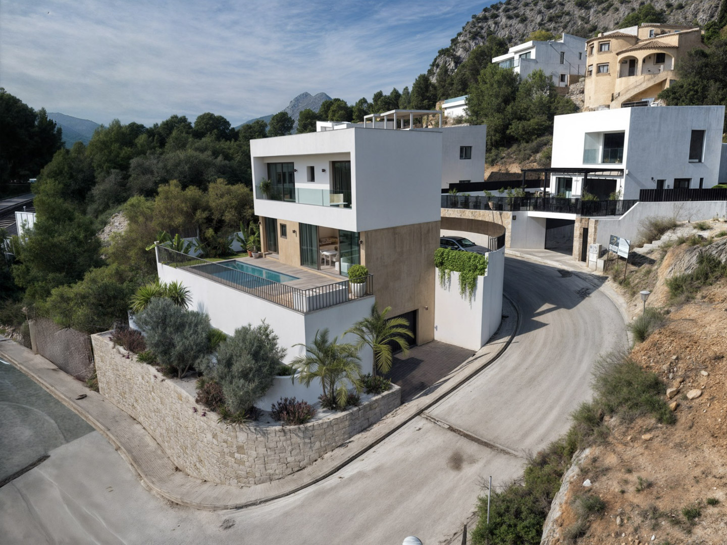 Vista exterior de la villa moderna con fachada blanca y niveles de piedra, destacando la terraza con piscina y el entorno natural de la Vall de Pop.