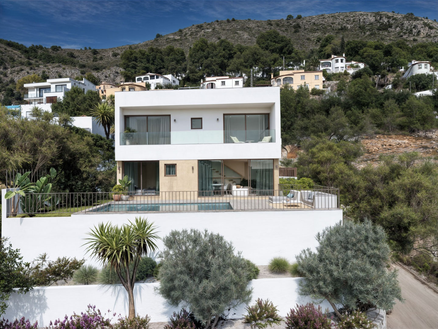 Vista exterior de la villa moderna con fachada blanca y ocre, destacando la piscina privada, amplios ventanales y terrazas con vistas a la montaña en Alcalalí.