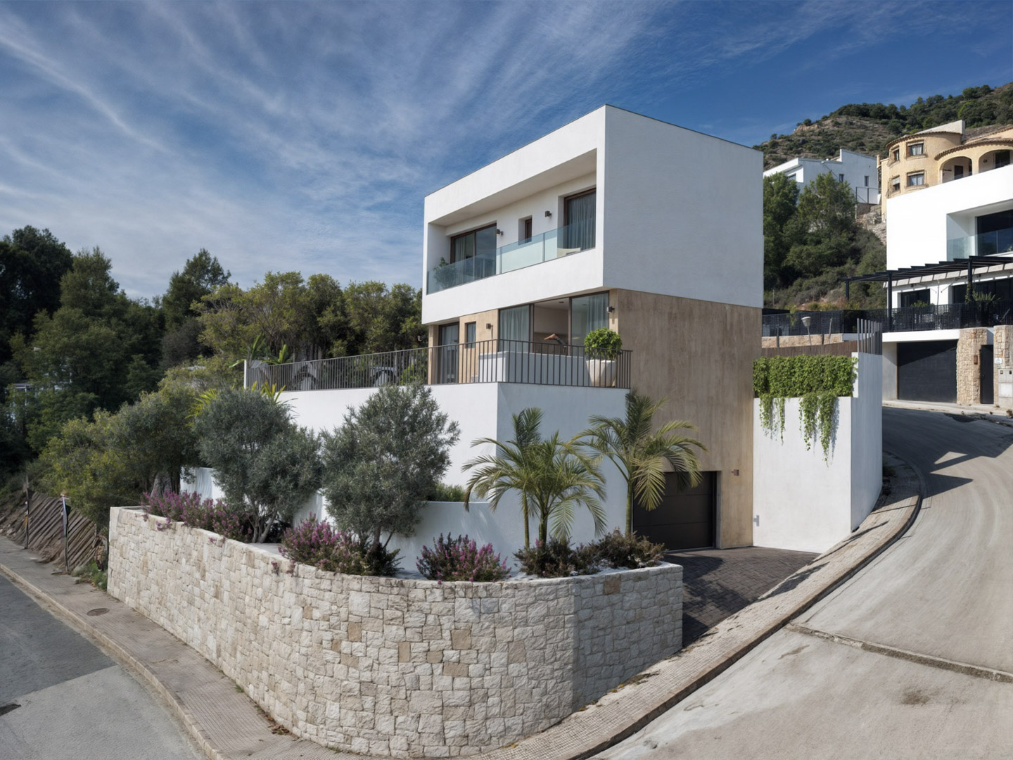 Vista exterior de una villa moderna en Alcalalí, con fachada de piedra natural, amplias terrazas con barandillas de cristal y garaje integrado en el semisótano.