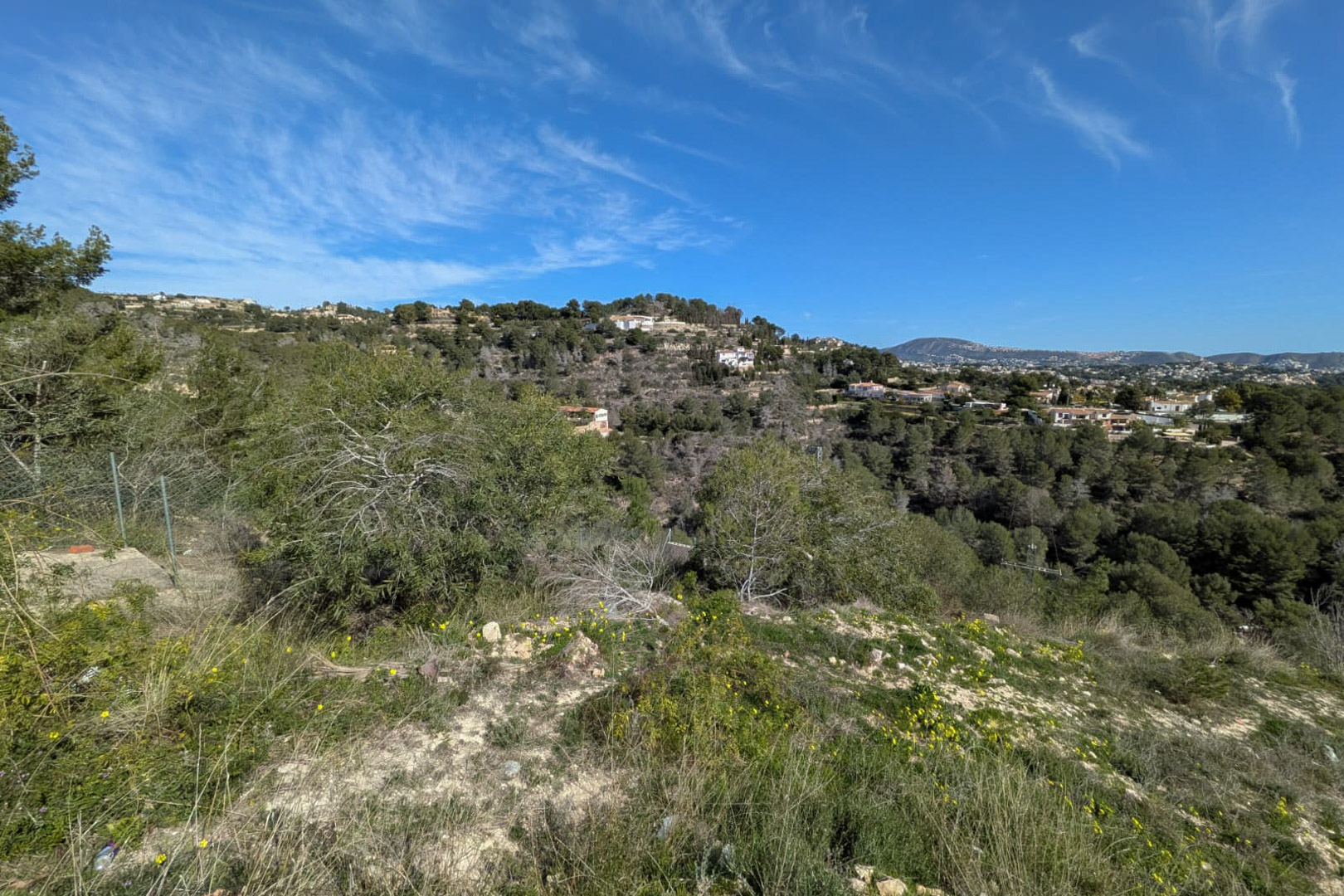 Vista elevada desde la parcela escarpada que muestra la vegetación natural y las colinas arboladas de Benissa bajo un cielo azul despejado.