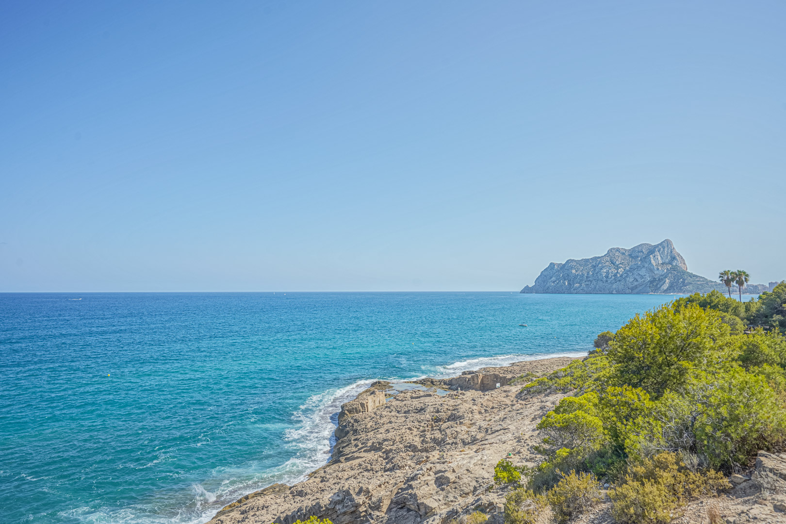 Vista panorámica de la costa rocosa de Benissa con el Peñón de Ifach al fondo, destacando las aguas turquesas y la vegetación mediterránea.