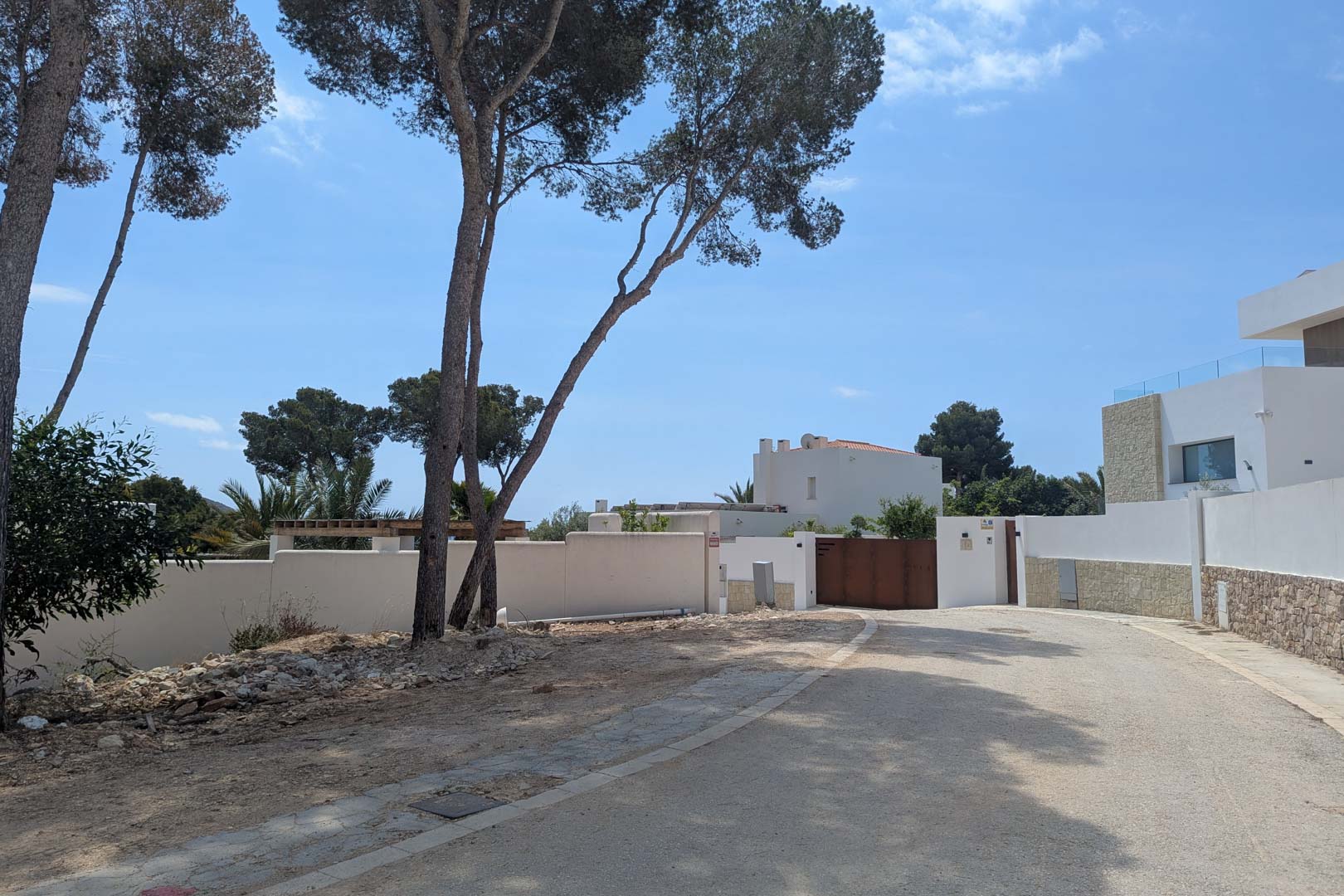 Acceso a la parcela en una zona residencial de Moraira, con muros perimetrales blancos, pinos maduros y calle pavimentada bajo un cielo despejado.