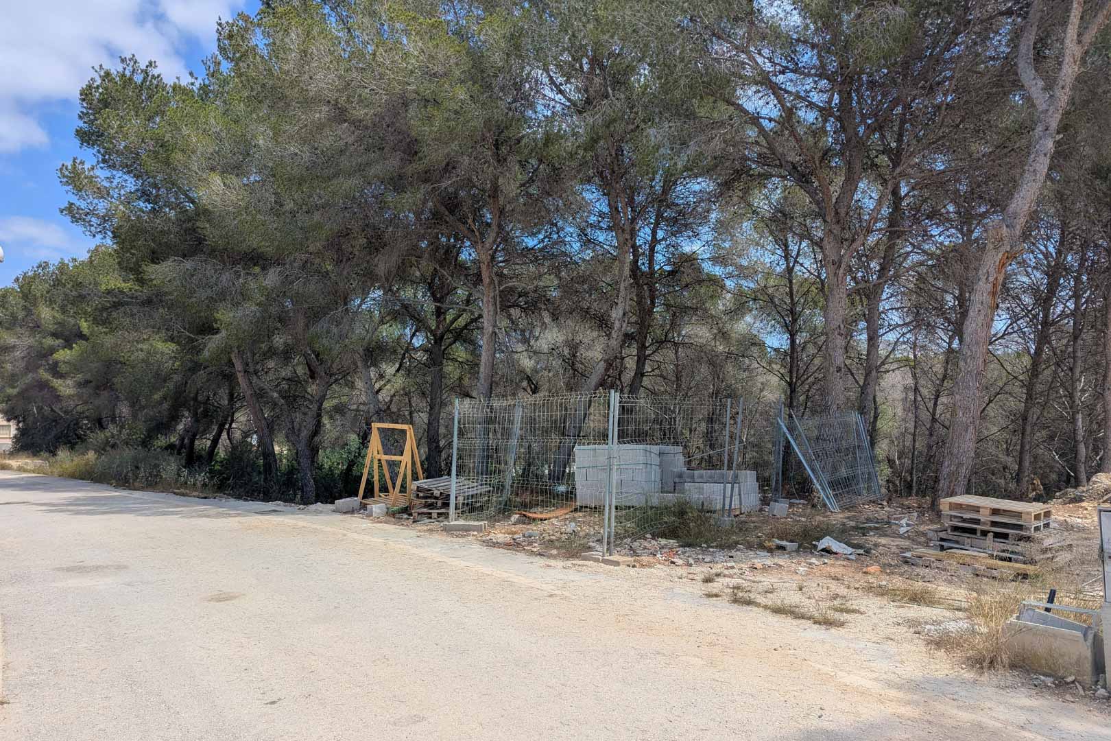 Parcela con pinos maduros y materiales de construcción iniciales junto a una calle asfaltada en Moraira, lista para comenzar la edificación de la villa.