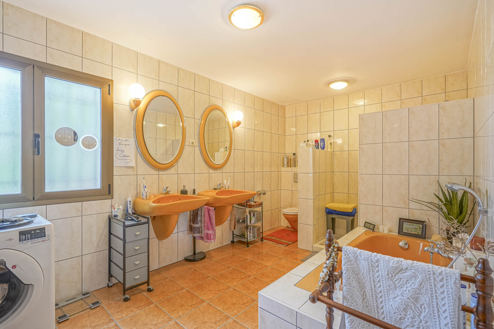 Cuarto de baño con lavabos naranjas y bañera a juego, baldosas color crema y ventana con luz natural.