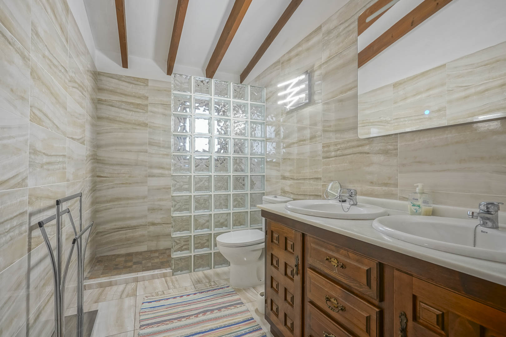 Cuarto de baño con pared de bloques de vidrio, tocador de madera con doble lavabo y azulejos veteados.