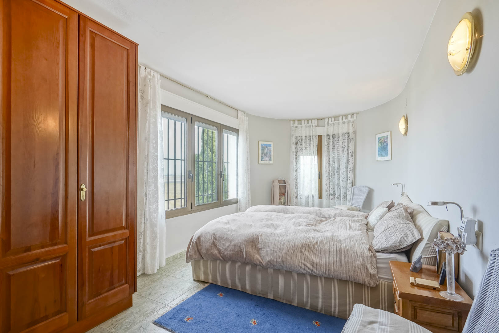 Dormitorio luminoso con armario de madera, ventanas con vistas y ropa de cama a rayas.