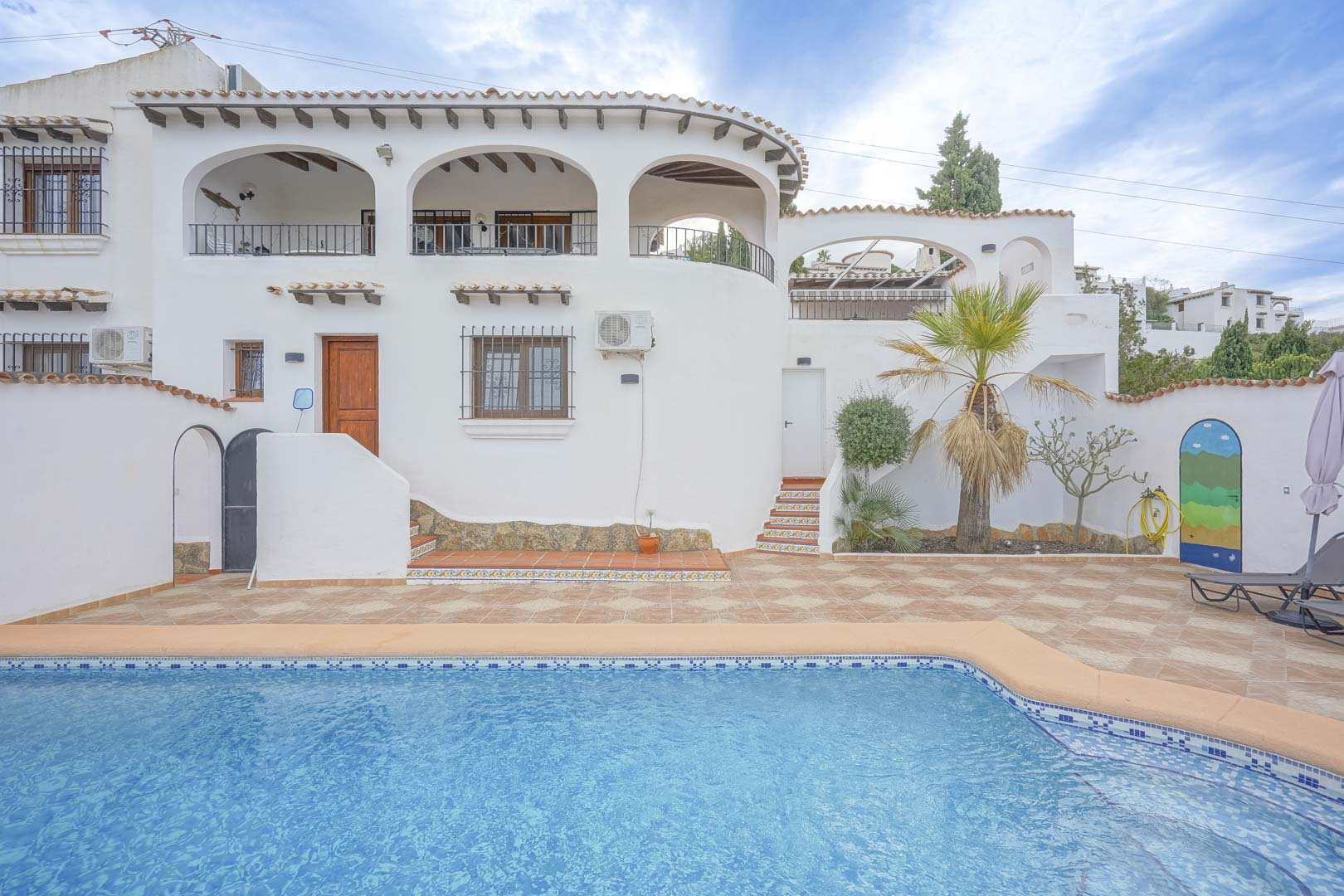 Vista de la fachada principal de la villa con piscina, terraza de baldosas y vegetación.
