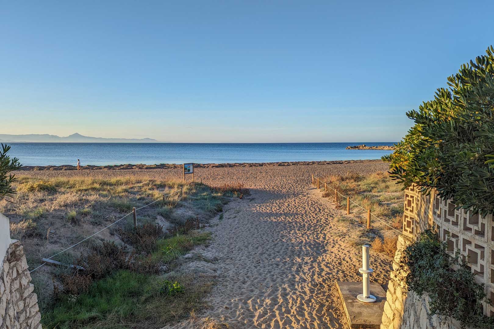 Vista de la playa arenosa con camino delimitado, vegetación costera y el mar azul bajo un cielo despejado.