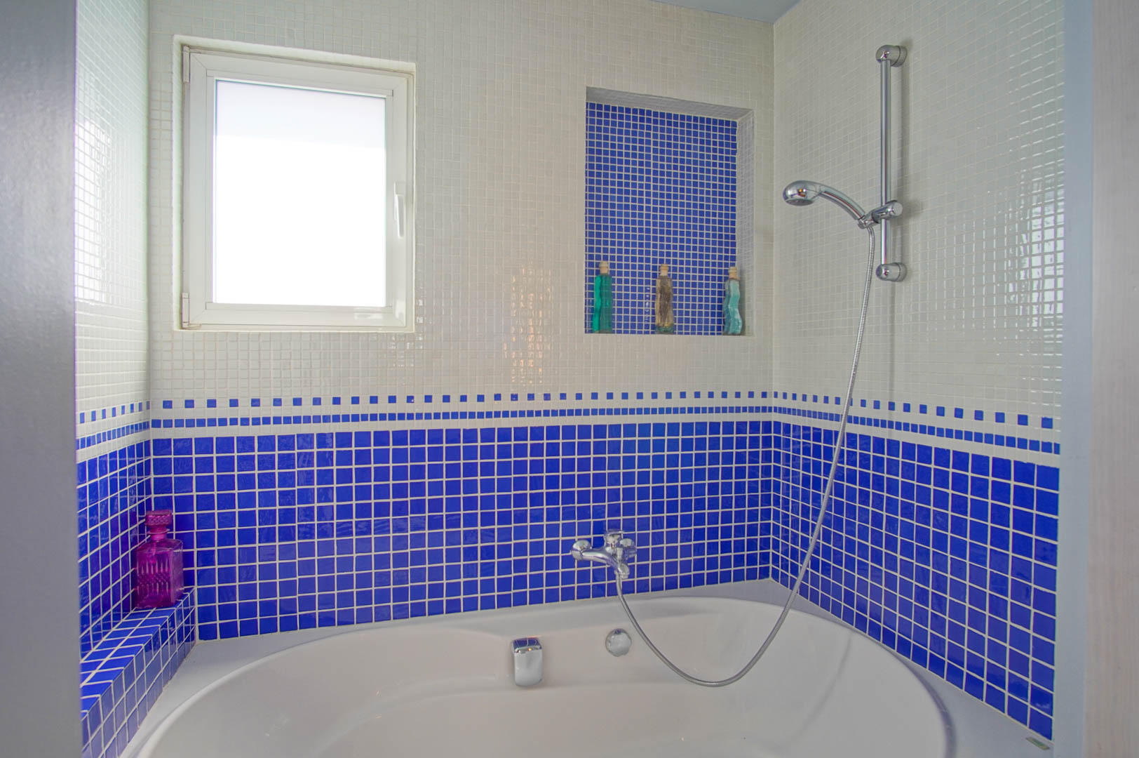 Baño con bañera ovalada y paredes revestidas en gresite blanco y azul cobalto. Incluye una hornacina integrada para accesorios y una ventana que aporta luz natural.