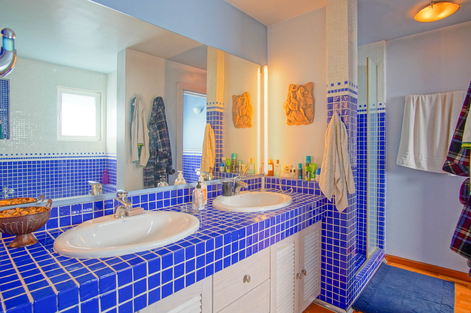 Baño con encimera de azulejos azules, doble lavabo y armarios blancos de lamas. Un gran espejo de pared refleja la zona de ducha y la iluminación cálida integrada.